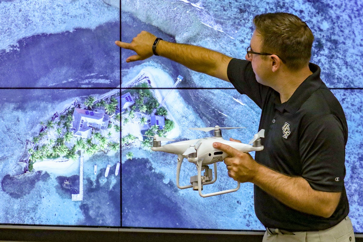 man wearing glasses and a black shirt points to a satellite image while holding a drone in his other hand