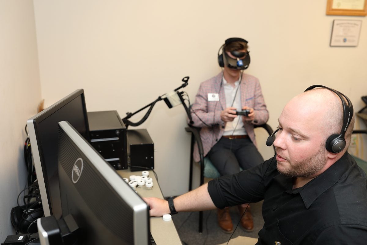 A bald man wearing headphones sits at a desk looking at a computer with another person sitting behind him wearing a VR headset