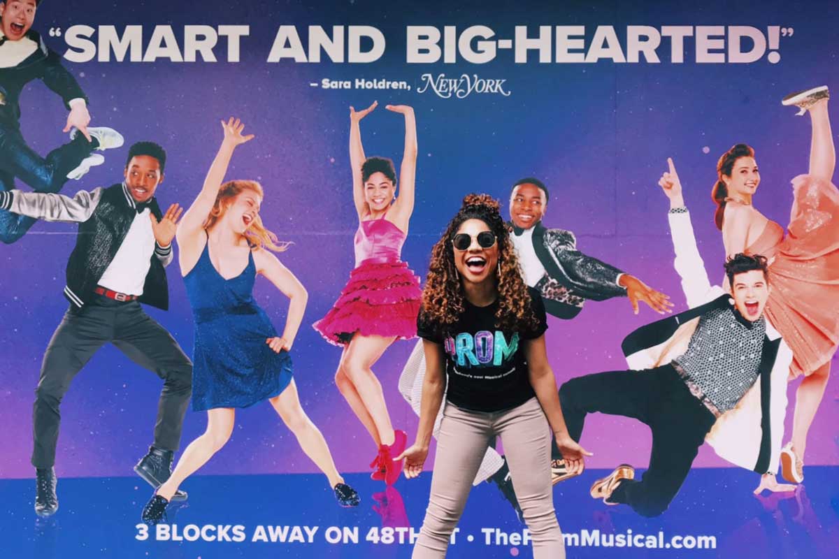 African-American woman wearing black shirt and khaki pants stands in front of billboard ad with text "Smart and Big-Hearted!"