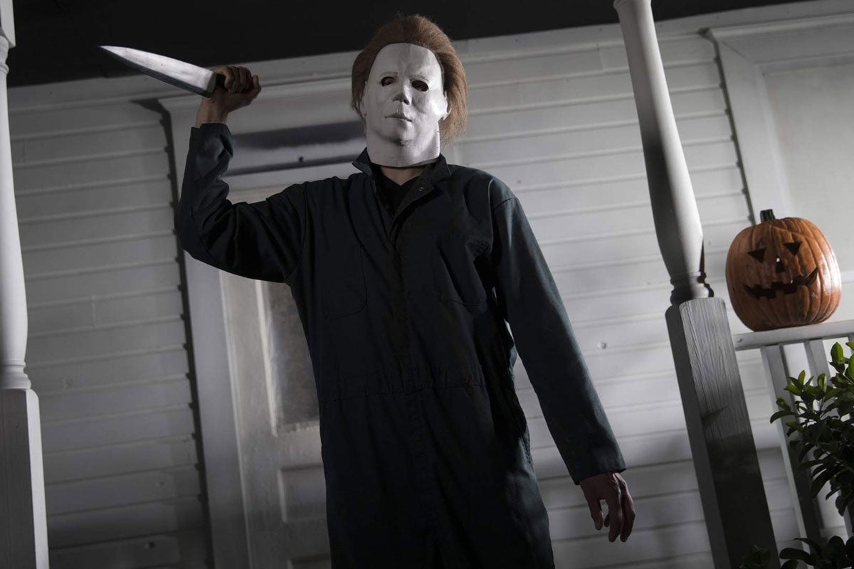 Michael Myers from the film Halloween holds a knife on a white front porch with a jack-o-lantern beside him