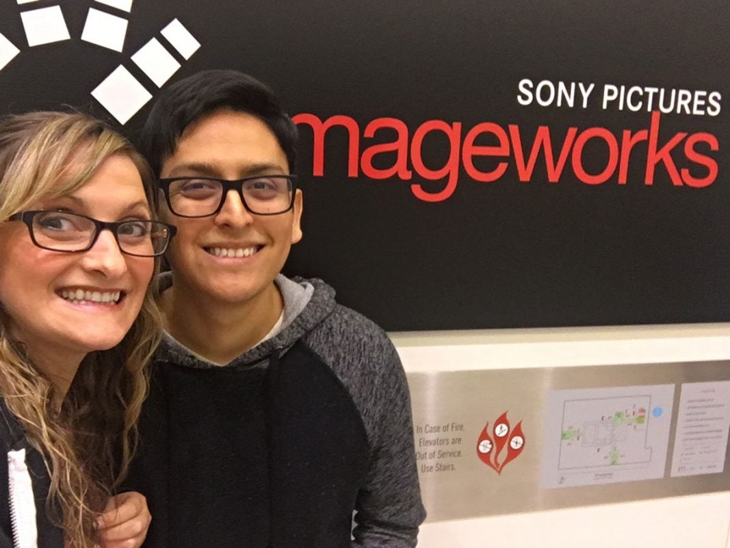 Young adult woman and man, both wearing black square frame glasses, pose in front of a red and black Imageworks sign