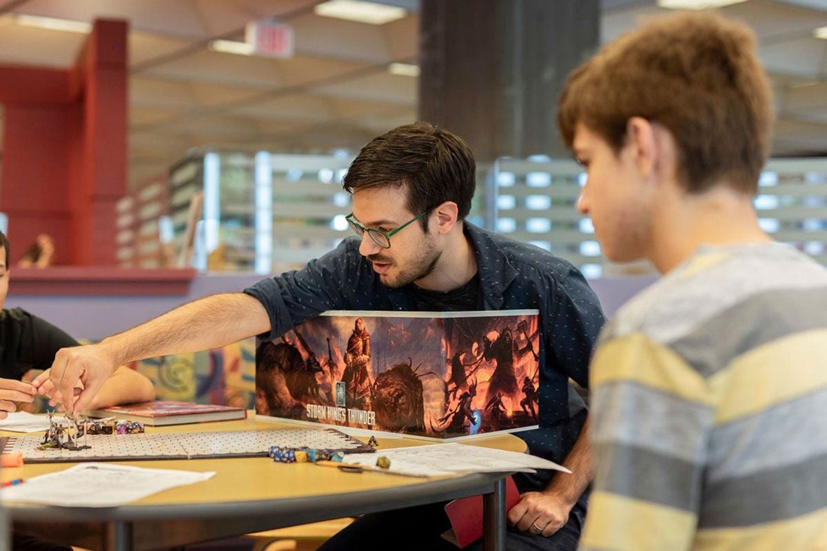 A man with dark hair and wearing glasses reaches across a round wood table with his right arm to move a piece on a game board as other players look on.