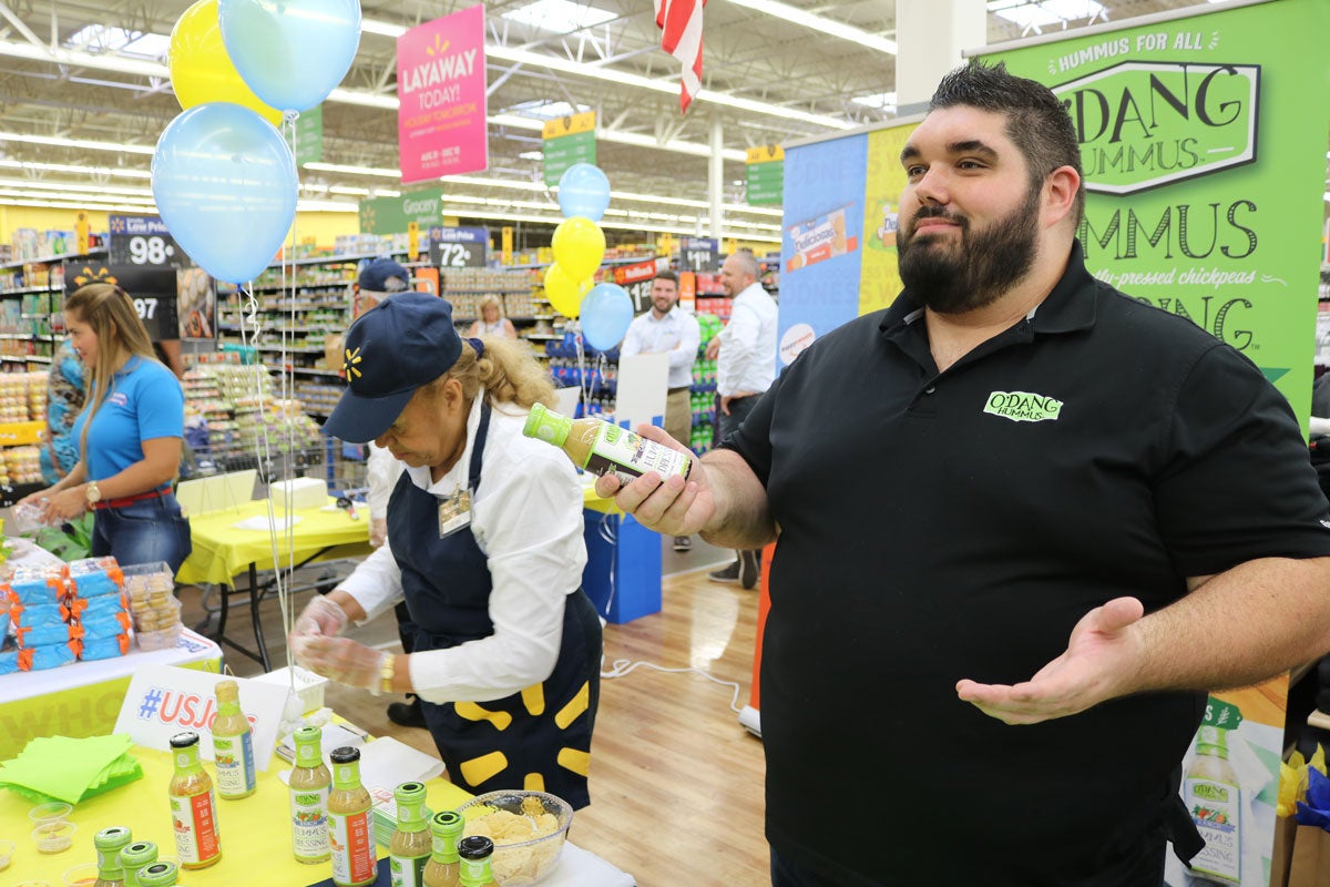 Man with dark hair and a dark beard wearing a black polo shirt stands in front of a table with green salad dressing bottles in a Walmart store