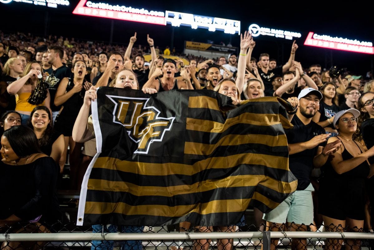 Fans cheering in the stands at night waving a UCF black and gold flag