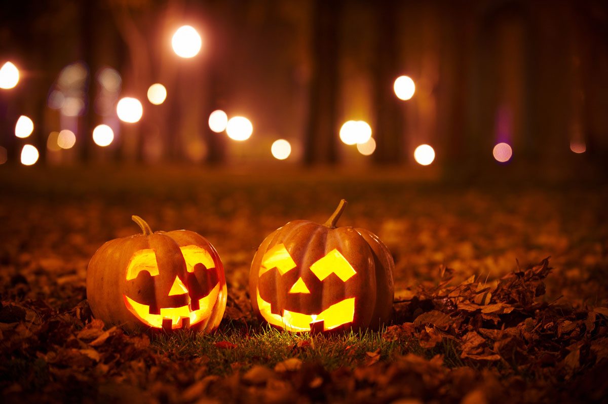 two lit-up jack-o-lanterns on the ground with trees and lights blurred in the background