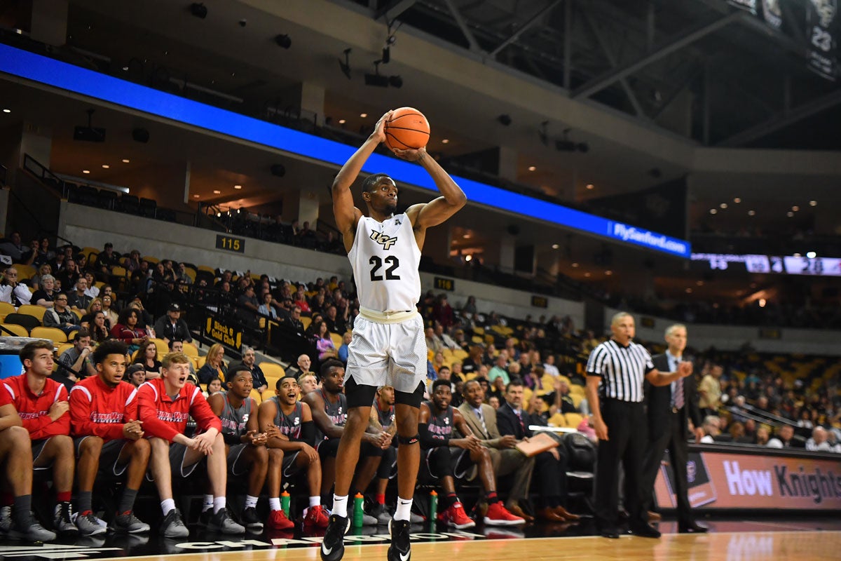UCF men's basketball player Chance McSpadden in a white uniform pulls up for a 3-point shot in front of opposing bench