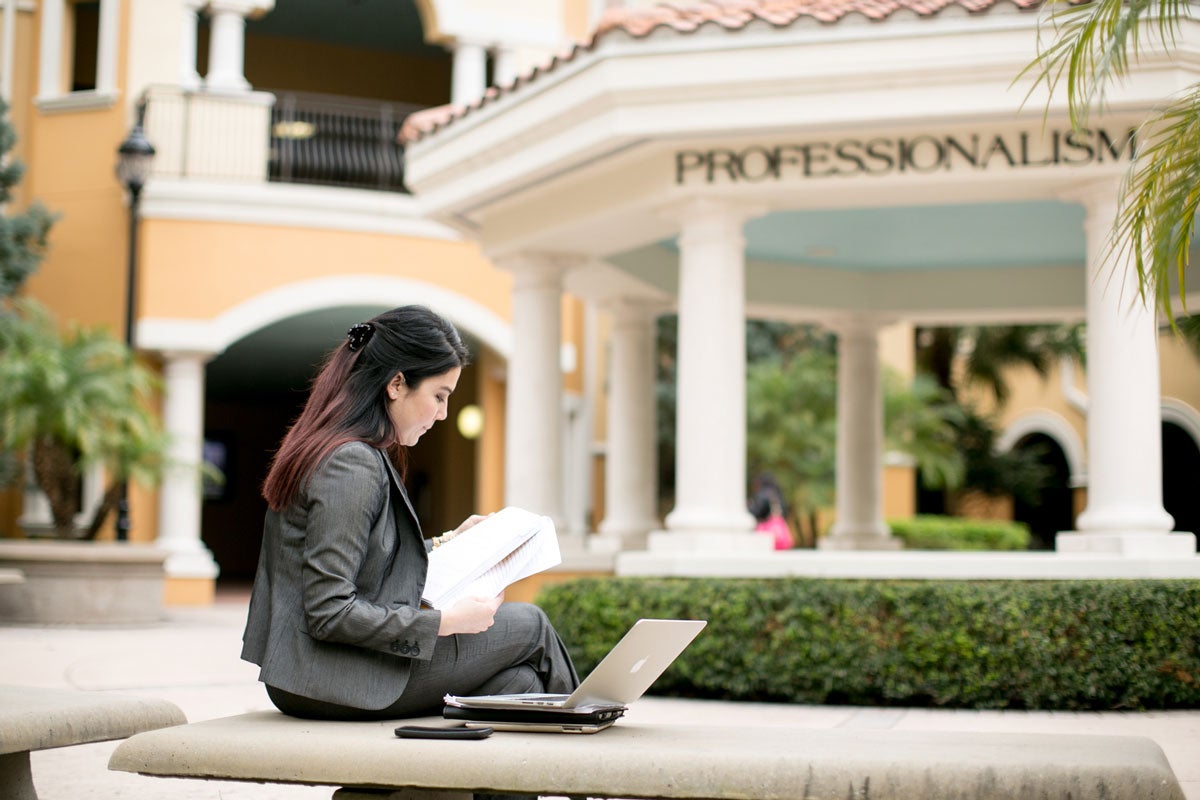 A woman with dark hair and wearing a gray business suit sits on a concrete bench with a laptop in front of a rotunda with white columns outside