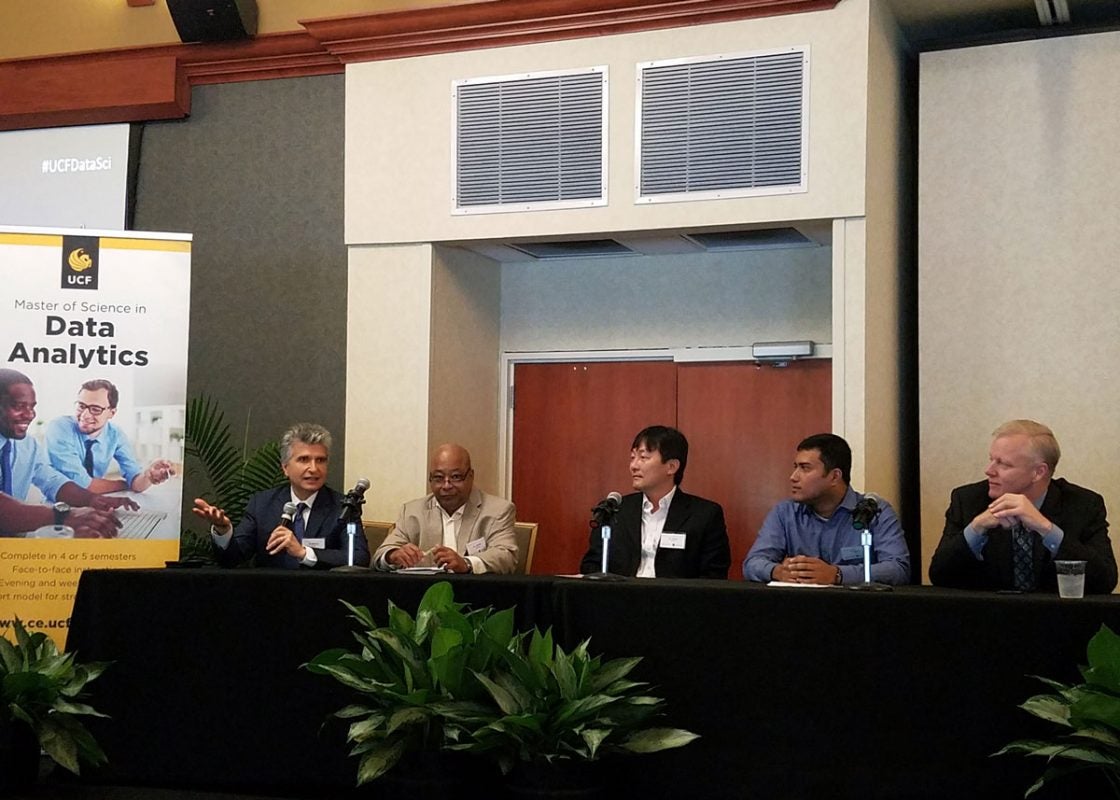 A panel of five men sitting at a table with green plants around it and a poster advertising UCF's data analytics program