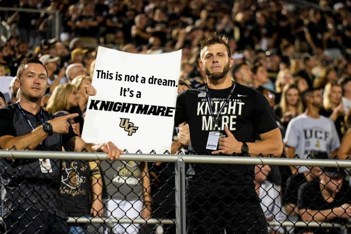 Two football fans wearing black t shirts hold a white sign that reads "This isn't a dream. It's the Knightmare"