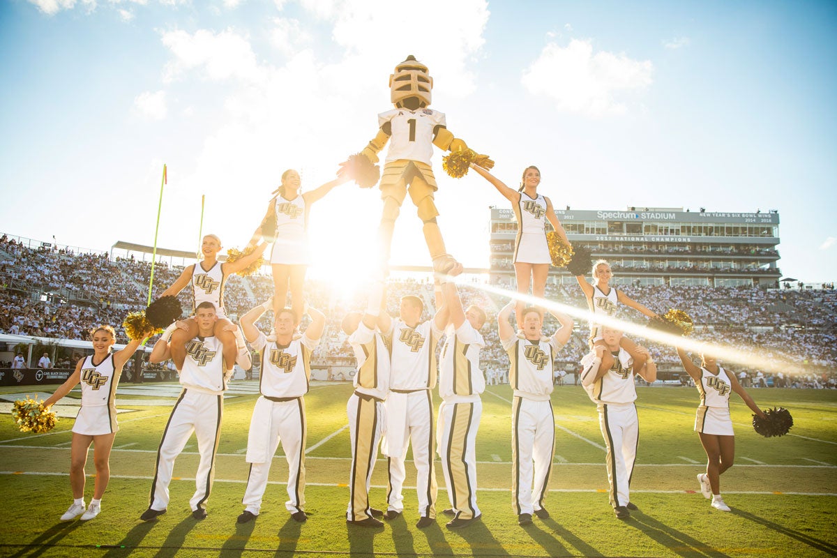 Knightro mascot stands atop a cheerleader pyramid on the football field with Spectrum Stadium crowd in the background as sun sets