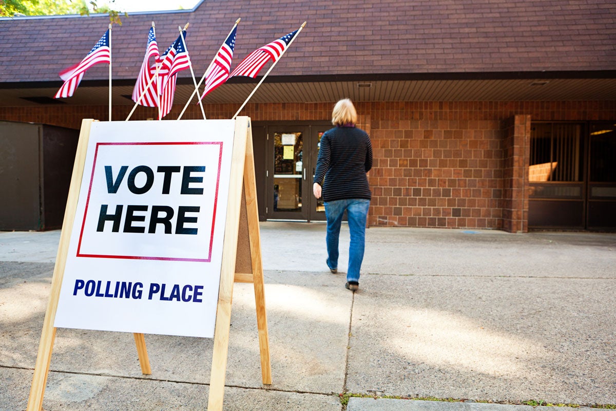 A blonde woman with a black long-sleeve shirt and jeans walks into a brick building with a sign outside that reads, Vote Here Polling Place