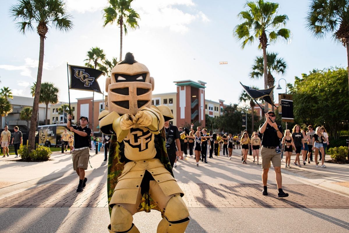 UCF mascot Knightro walks down the road with UCF cheerleaders and flags waving behind him