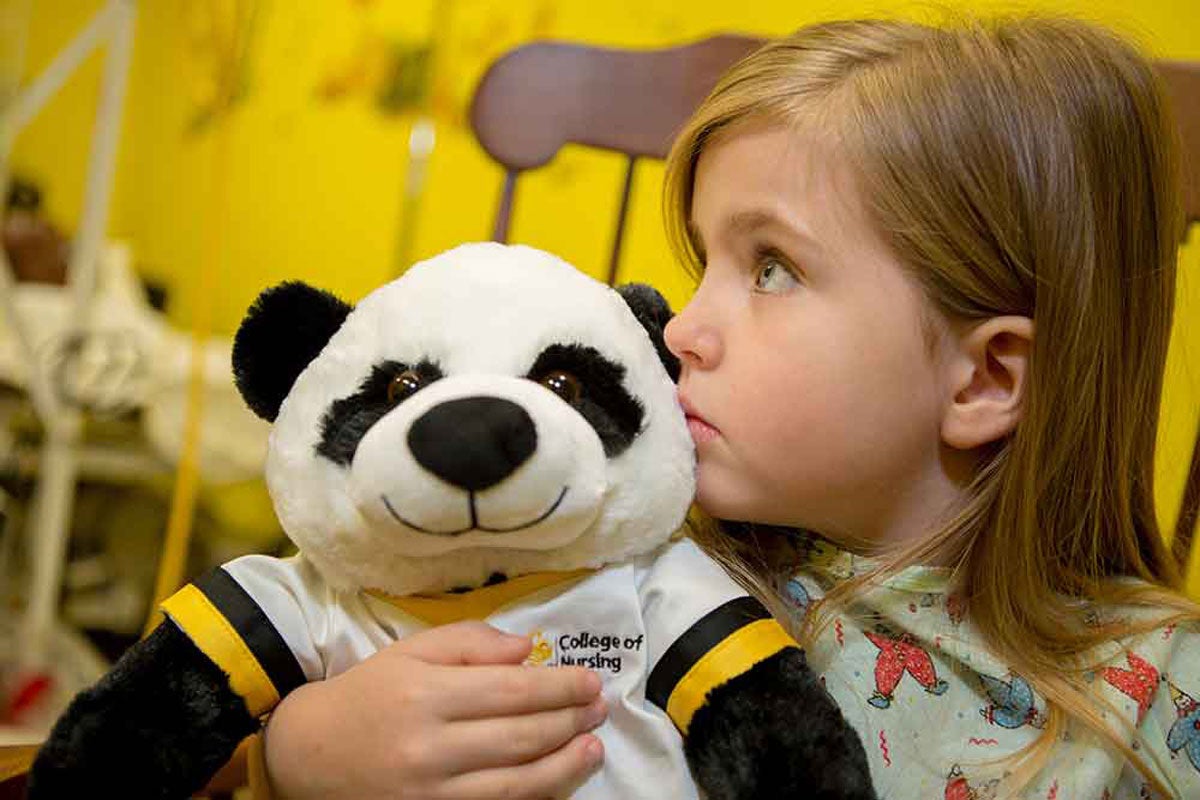 a girl toddler with blonde hair clutches a stuffed panda wearing black and gold nursing scrubs