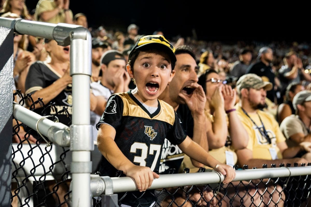 a young fan wearing a black and gold football jersey cheers in the stands
