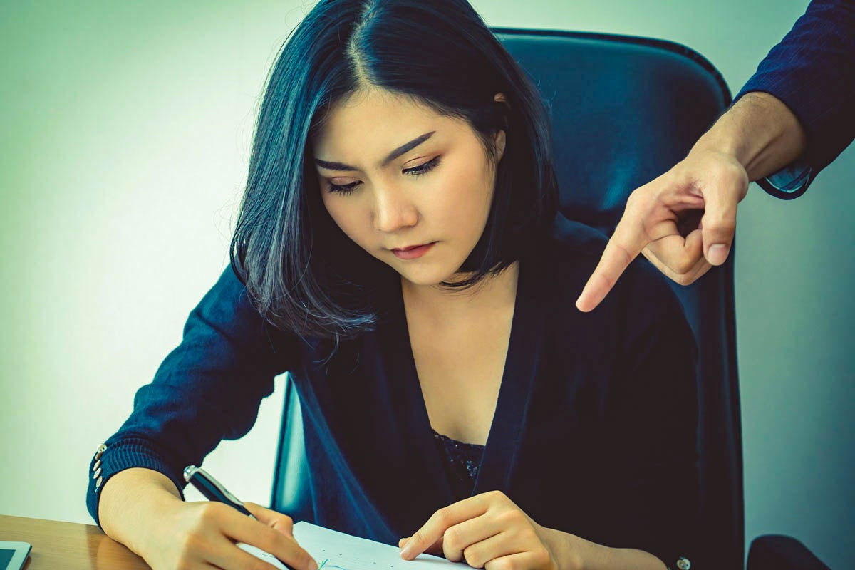 A dark-haired woman sits at a desk with a pen in hand as another's hand points over her shoulder at her paper