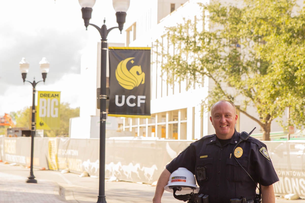 A bald man wearing a police uniform stands outside in front of a light pole with a UCF banner and building in the background