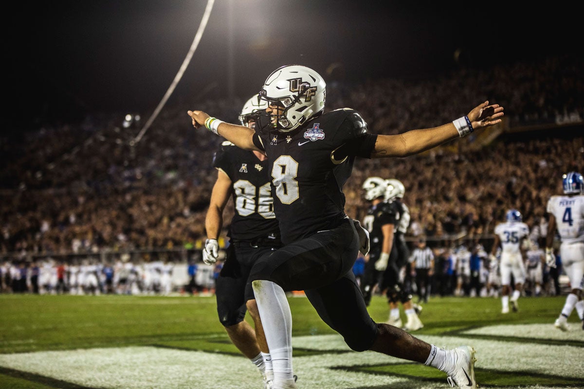 A football player wearing a black uniform and white helmet runs in the end zone with his arms extended like wings