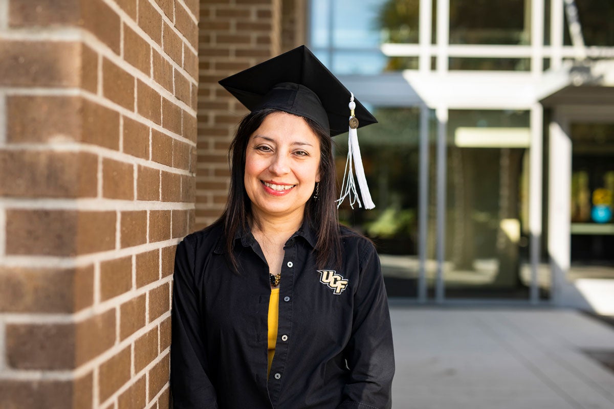 Woman with dark hair and black long sleeve shirt with UCF logo poses in black graduation cap and black and white tassel next to a brick wall