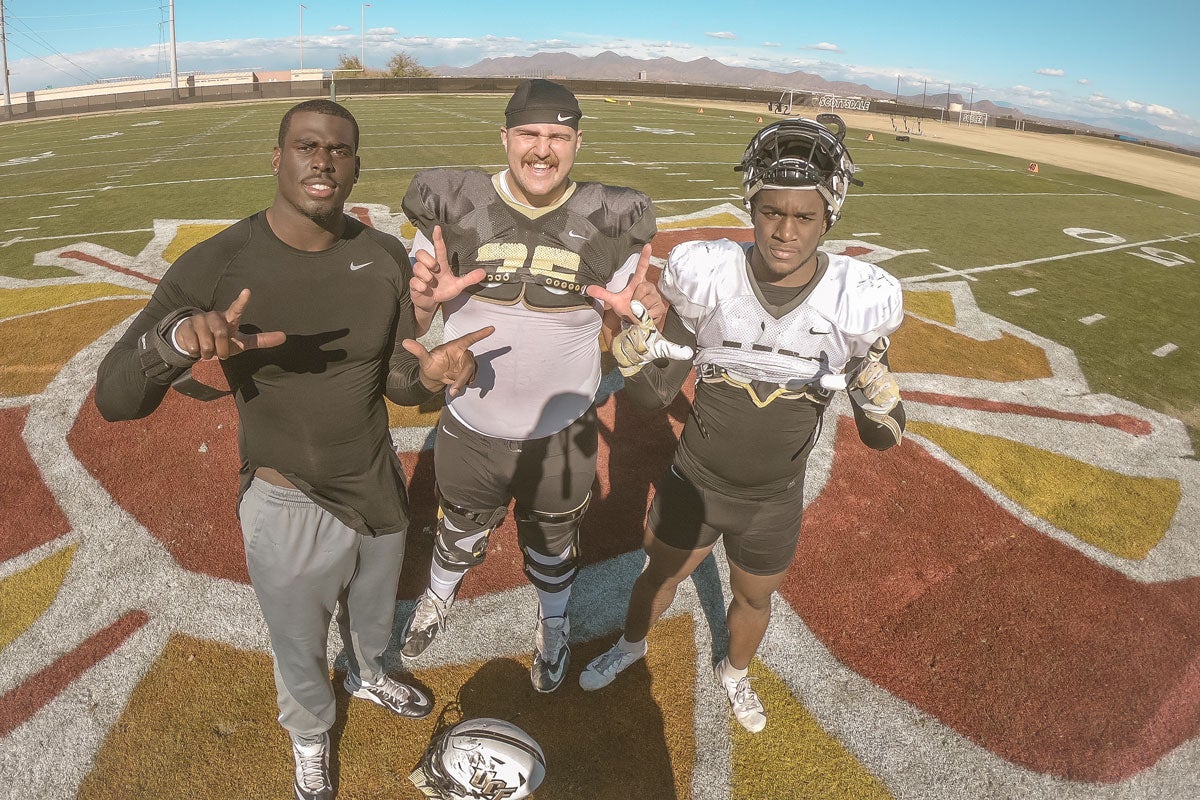 Three UCF football players stand on football field making L shapes with their hands on a sunny day
