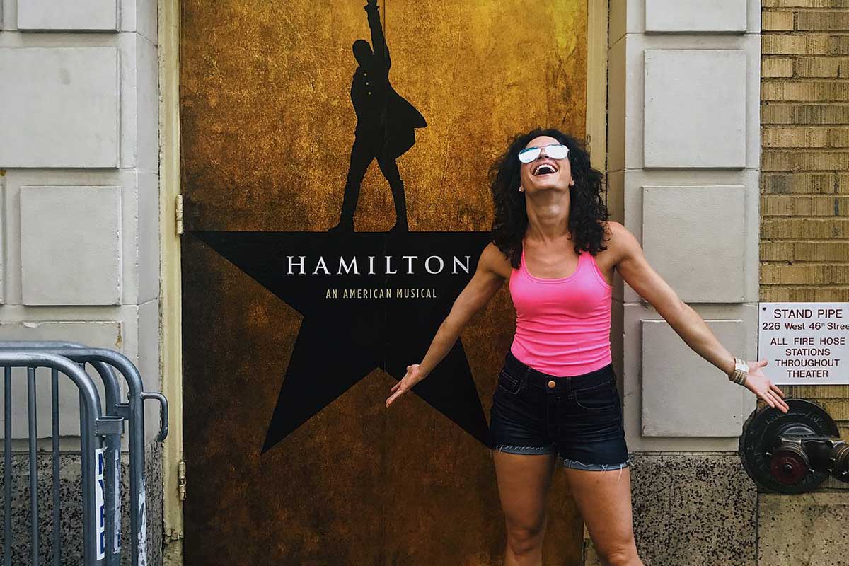 Brunette woman wearing pink tank top stands in front of Hamilton poster