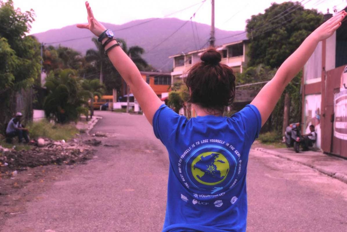 A girl with brown hair wearing a blue T shirt holds her arms up in a V shape on a gravel road with mountains in the background