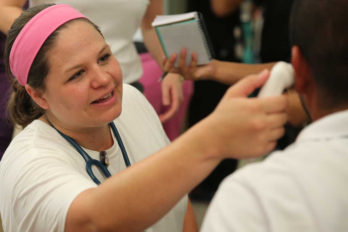 A woman with a pink head band and white t shirt checks someone's temperature with an ear thermometer