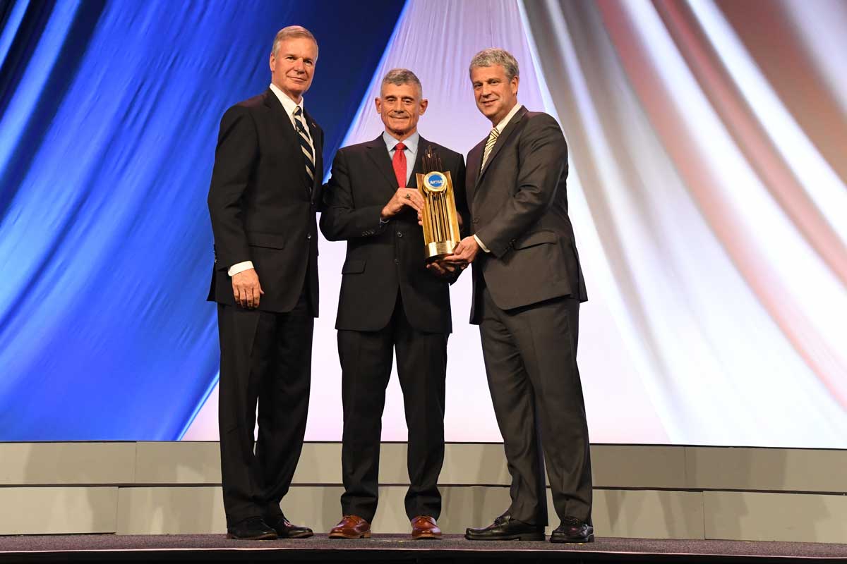 Three men on stage as one hands the middle man a gold trophy