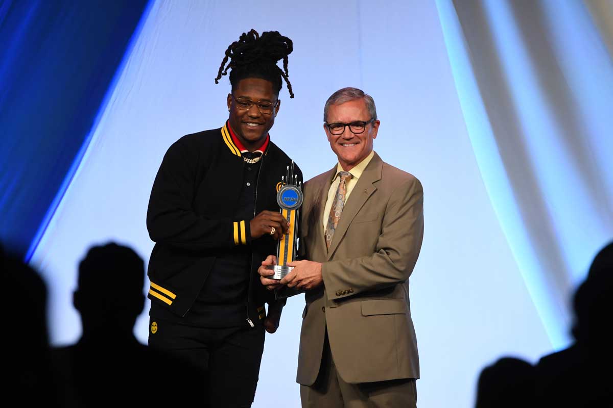 Dale Whittaker, wearing tan suit, presents gold trophy to Shaquem Griffin, wearing a black jacket with yellow cuffs