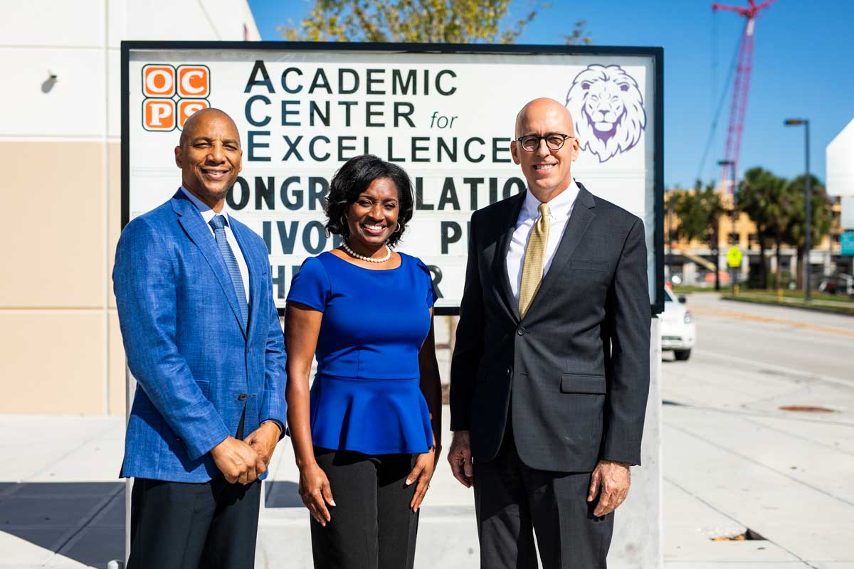 Two men and a woman stand in front of an Academic Center for Excellence sign outside on a sunny day