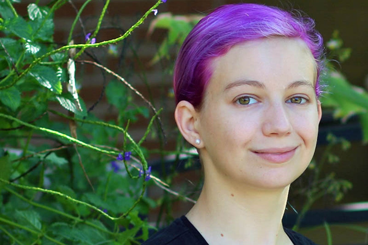 White female with purple hair poses in front of shrubbery