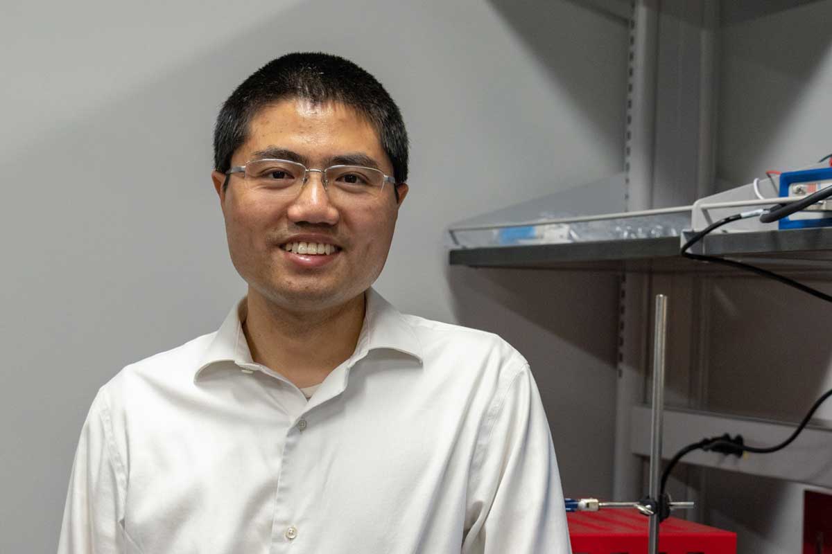 Asian man wearing glasses and white collared shirt stands next to a metal shelf