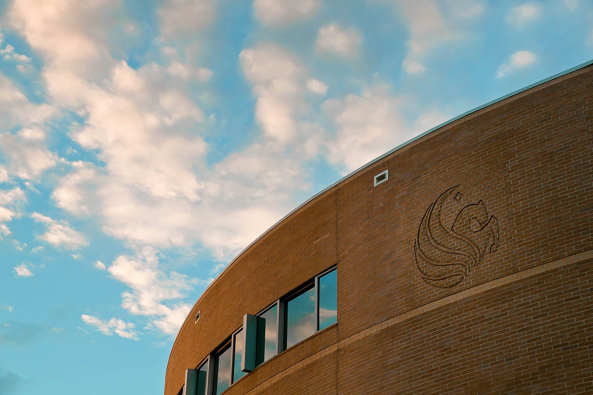 A campus building with a Pegasus logo and blue sky and clouds above.
