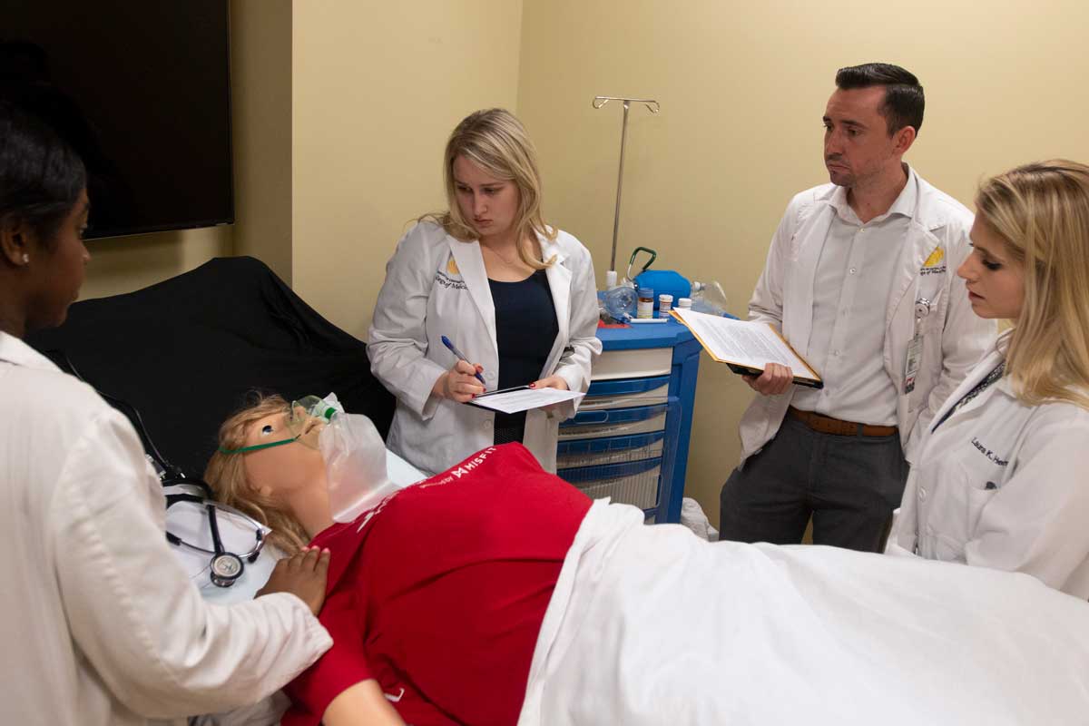 Four medical students stand around a mannequin patient's bedside