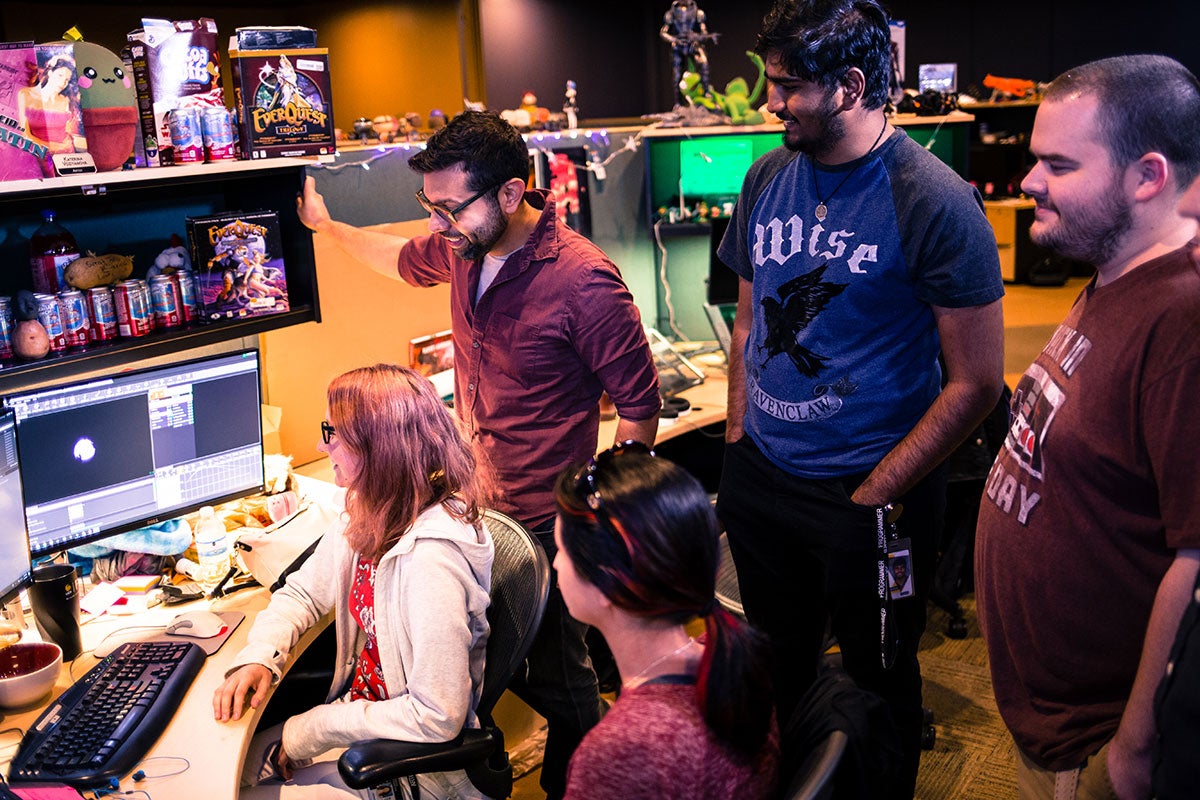 Four people crowd around one woman sitting in a chair working on a computer.