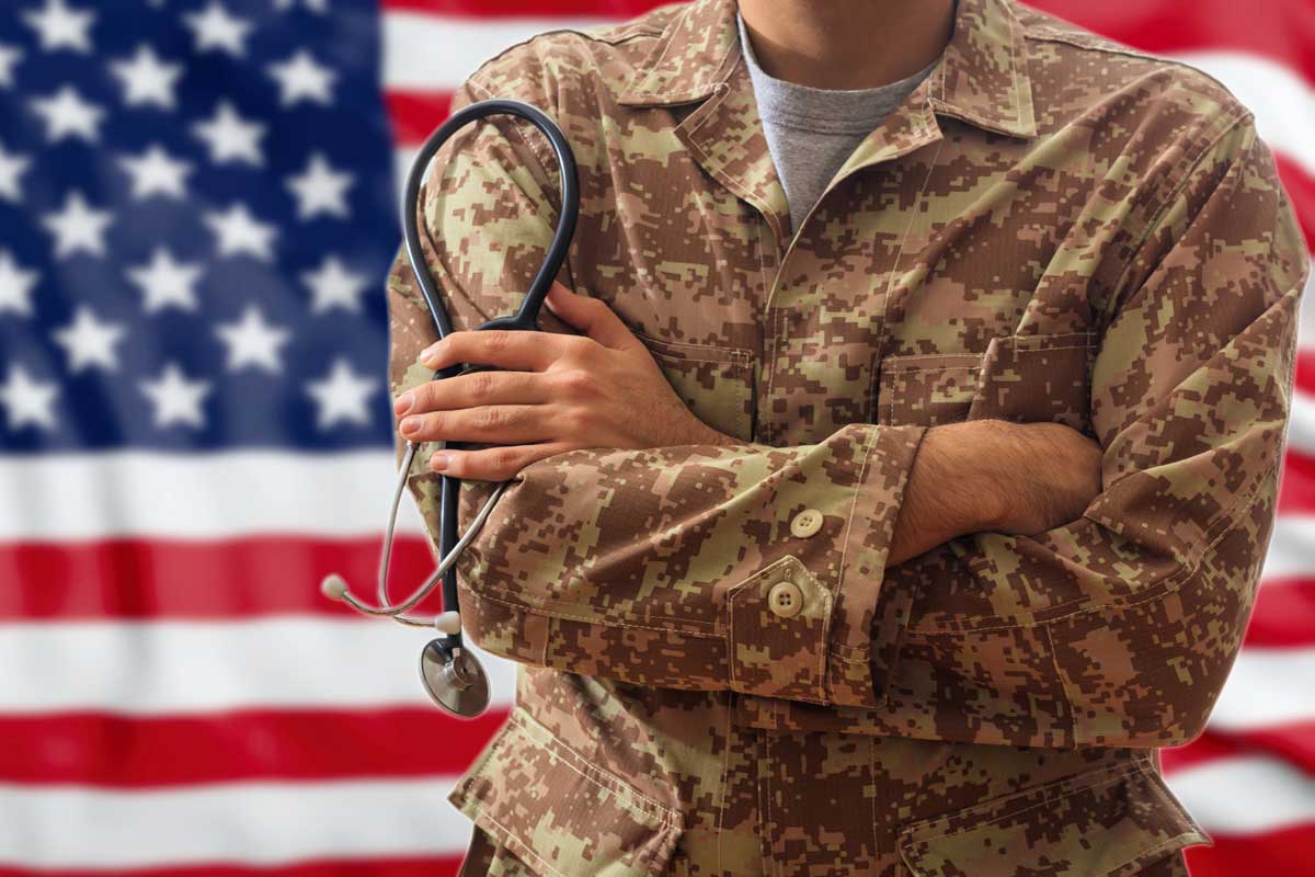 Person outfitted in camouflage jacket with arms crossed stands in front of American flag