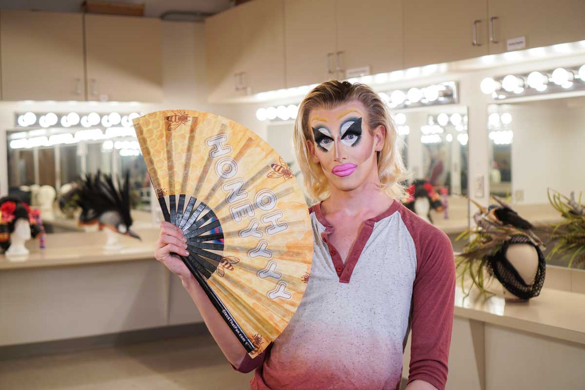 Man with blonde hair and drag makeup holds yellow fan in makeup room