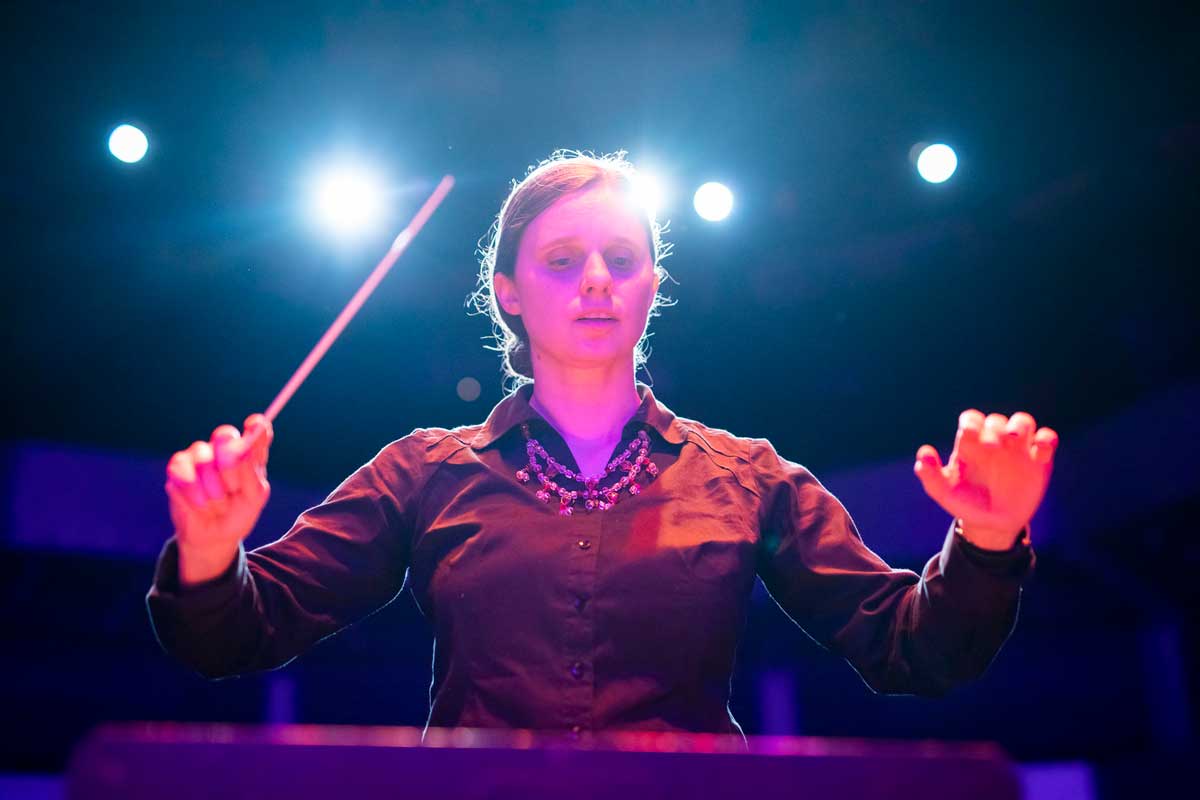 Woman in black long sleeve shirt holds conducting baton on stage with lights behind her