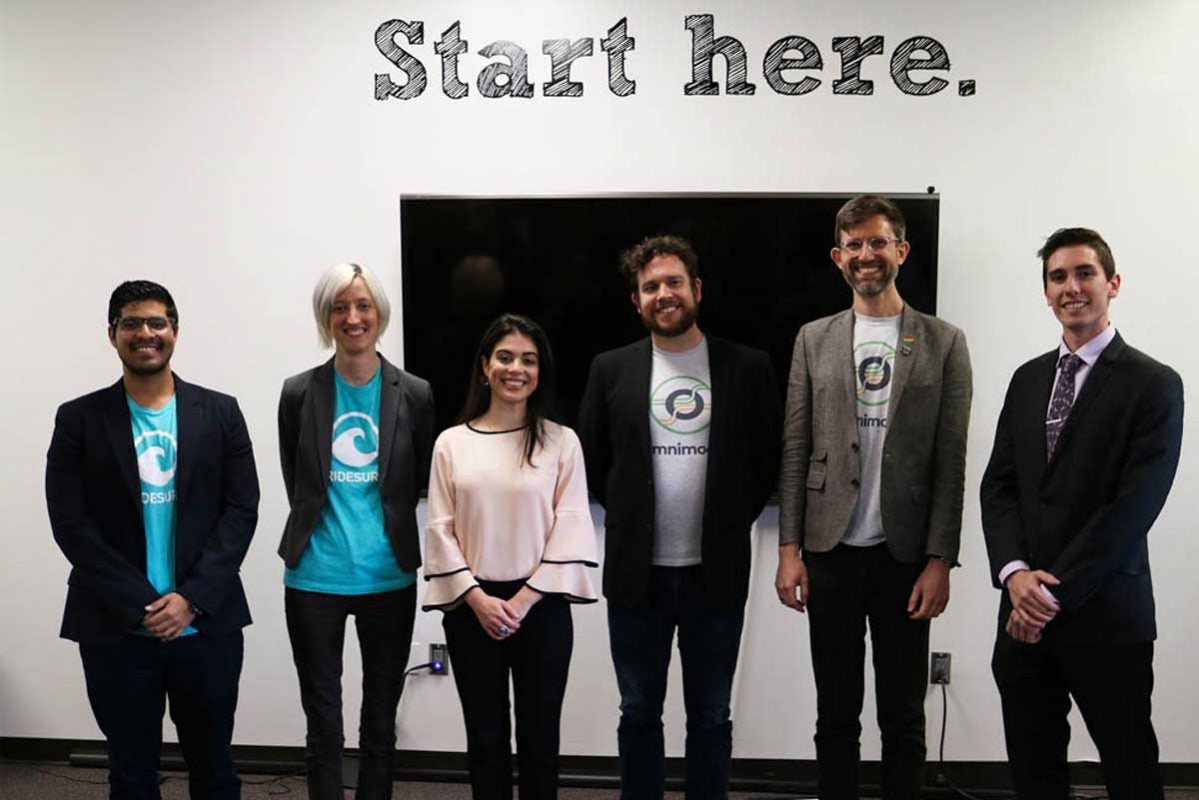 Six individuals stand in front of white wall with decal: Start here.