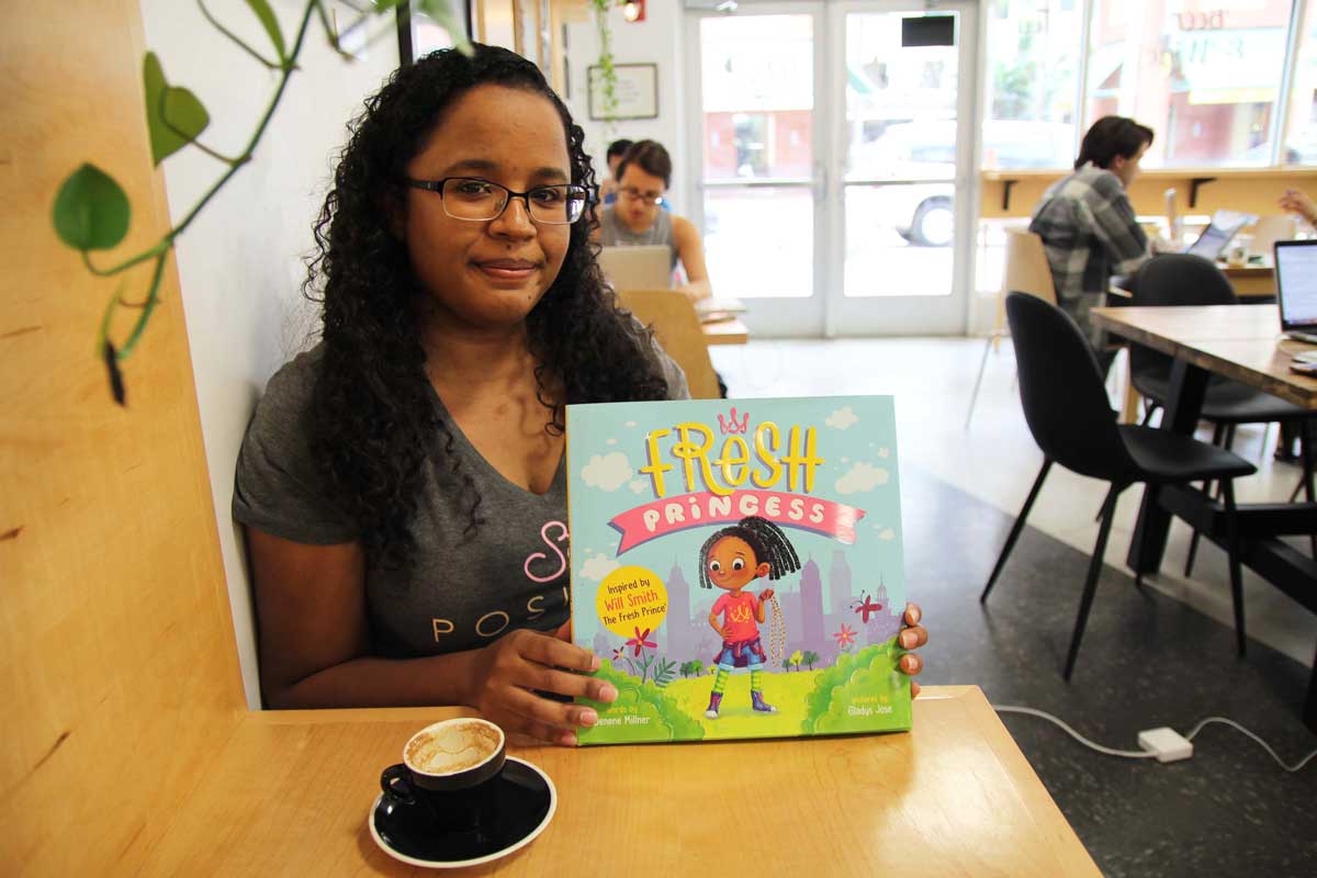 Woman with dark, long hair, sits at a wooden table with a coffee mug and holds up children's book