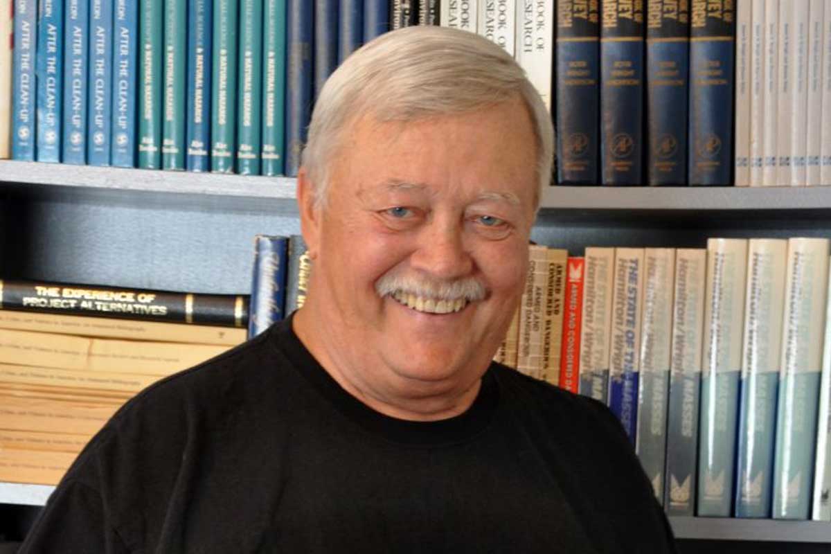 Man with gray hair and mustache stands in front of book shelf