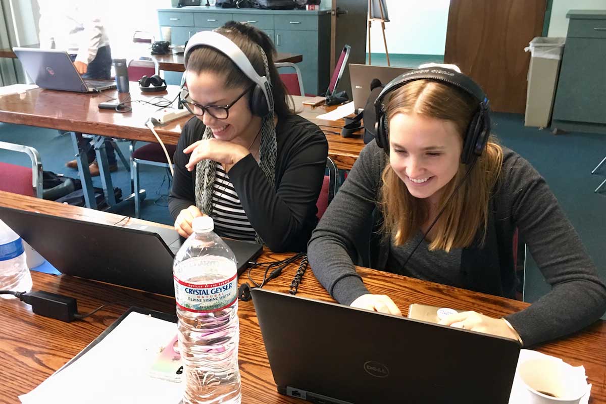 Two women with headphones on sit at a desk in front of laptops