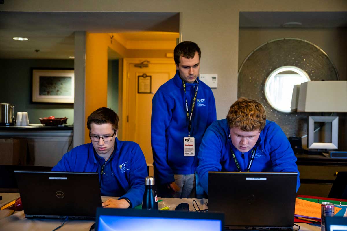 three men stand at a desk of computers