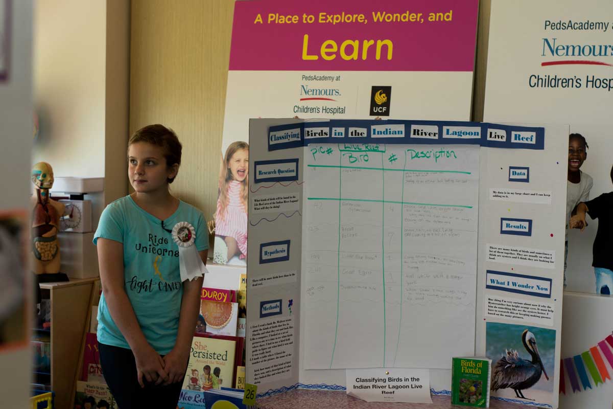 Young girl wearing a blue shirt stands next to science board display