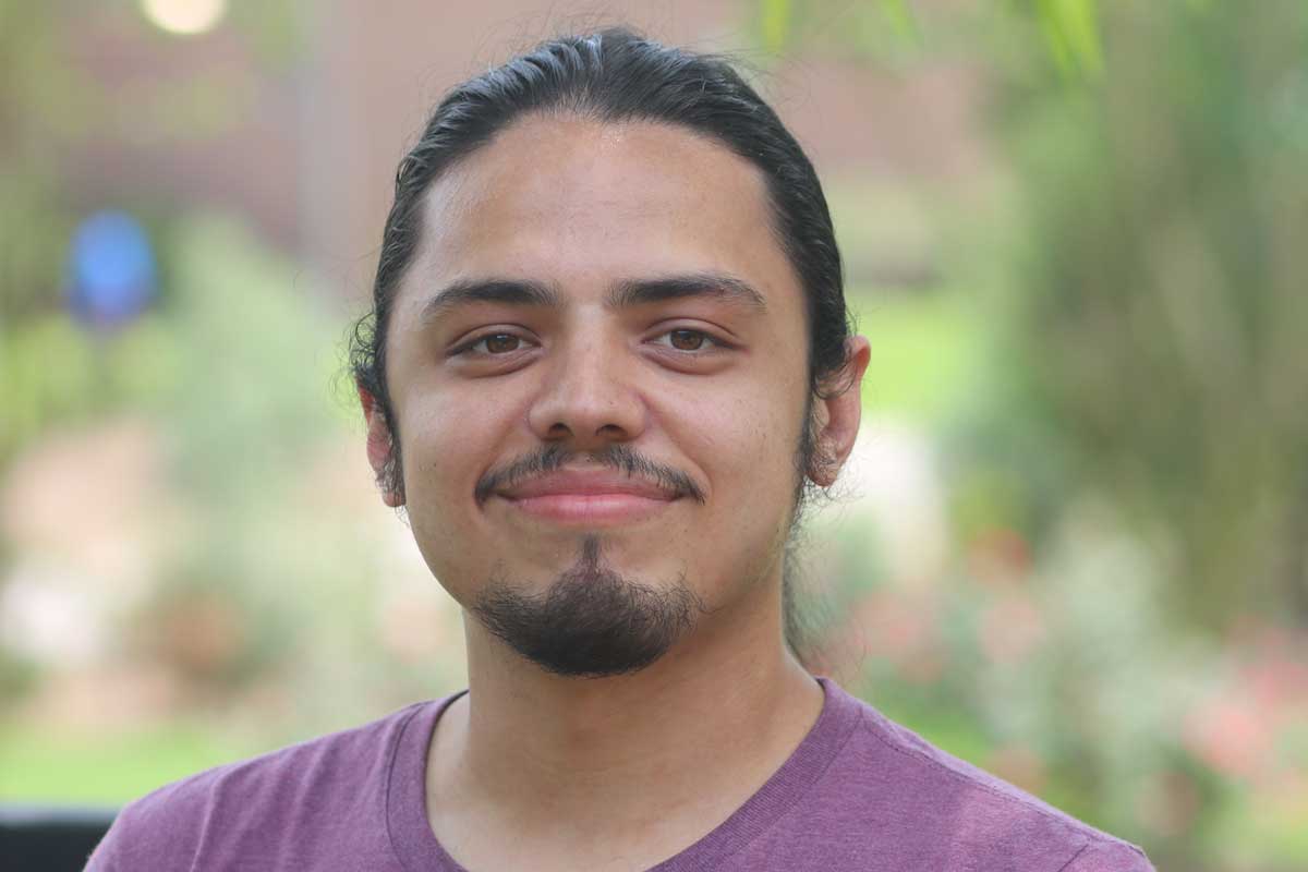 Man with dark hair and goatee smiles in front of greenery