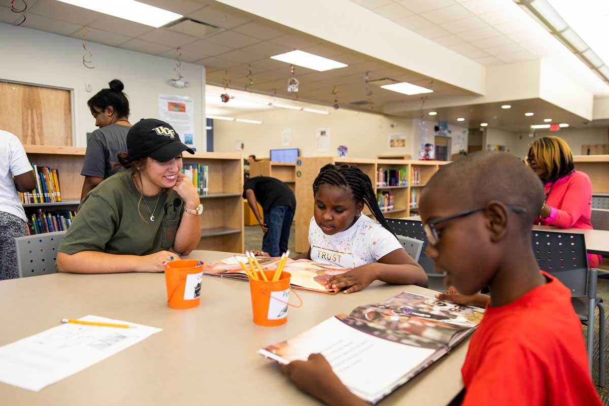 College student wearing a baseball cap and green shirt sits with two children reading at a table