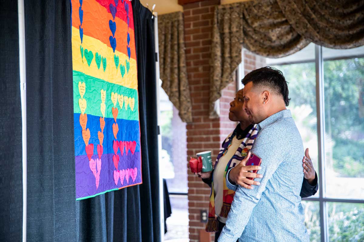 Two people stand in front of a rainbow colored quilt with L-O-V-E stitched in hearts