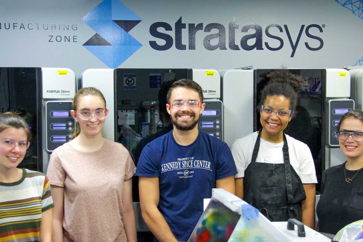 Five people stand in lab under the sign Stratasys sign