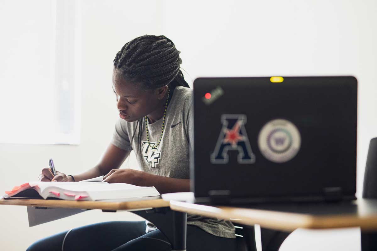 Female wearing gray UCF shirt sits at a desk with an open book and pen and a laptop