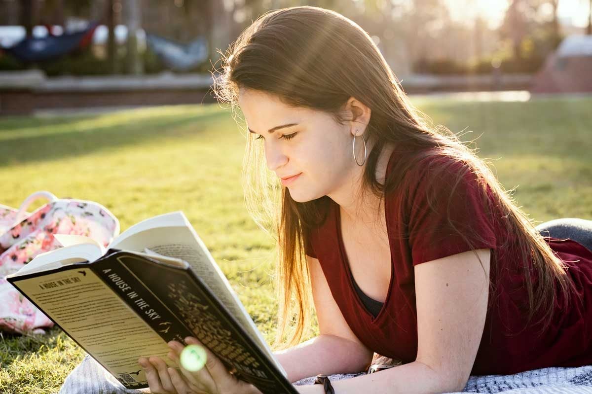 Young woman lays on stomach reading a book outside on the grass