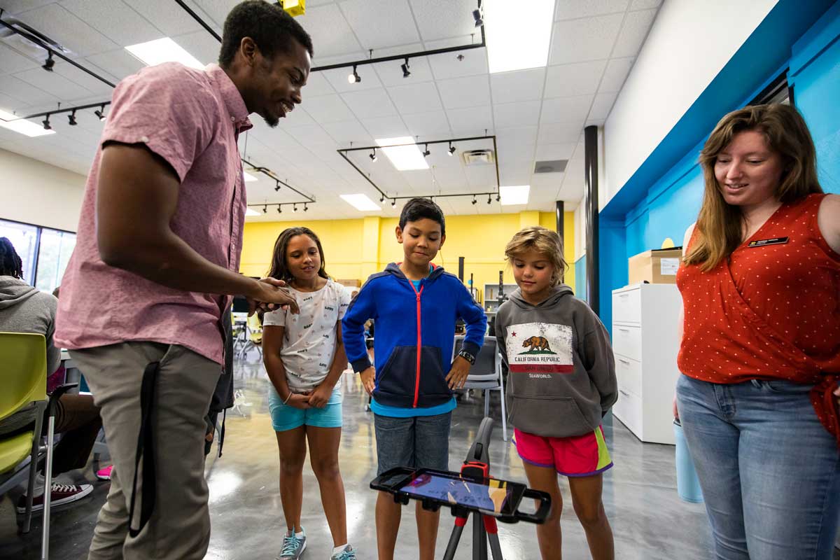 Two grad students observe three children working on an iPad and tripod in classroom